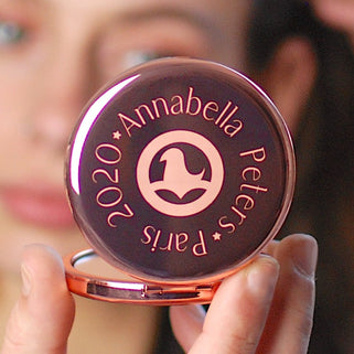 Rose gold compact mirror with personalised logo in the centre and text engraved around the outside, shown in close up with model.