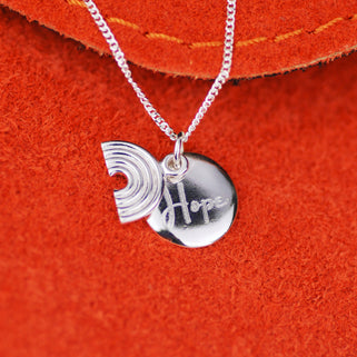 Sterling silver rainbow necklace with personalised circle disc engraved with "Hope" shown in close up shot on red background.